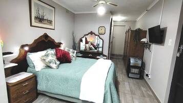 A bedroom with a bed, a bedside table, a mirror, a ceiling fan, a clock, a television, and a wardrobe.