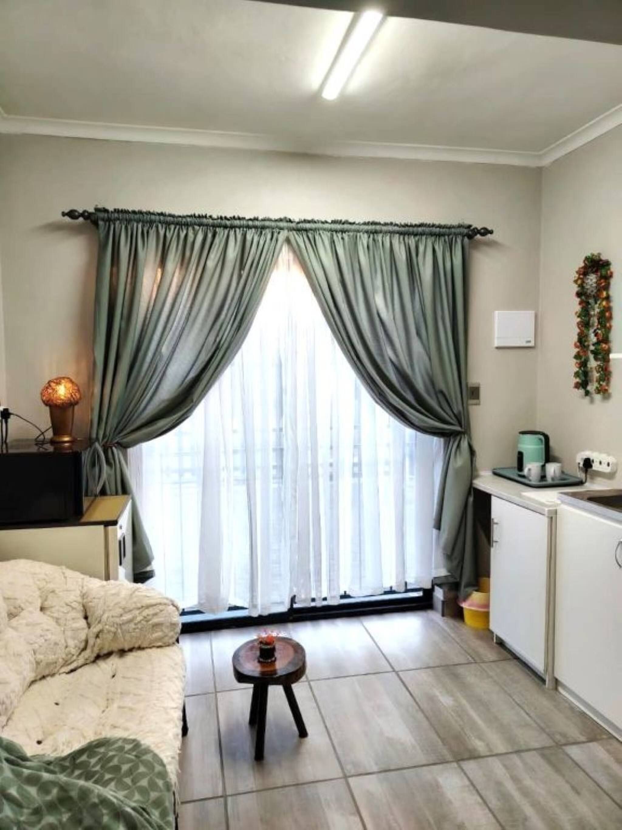 A small, well-lit room with a bed, a table, a window with curtains, a radiator, a wall-mounted light, and a small kitchenette area.