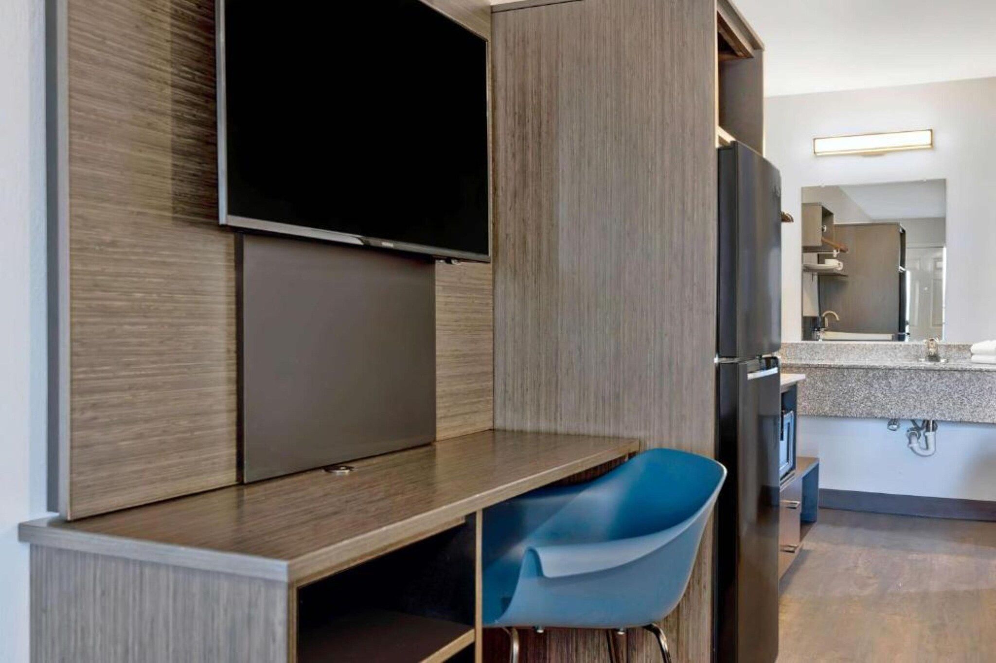 A modern hotel room with a flat-screen TV mounted on the wall, a built-in kitchen area with a countertop and sink, and a blue chair.