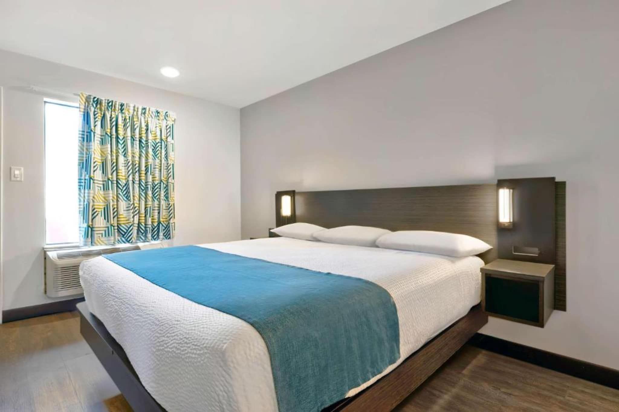 A modern hotel room with a large bed, a window with patterned curtains, a bedside table with a lamp, and a wall-mounted light fixture.