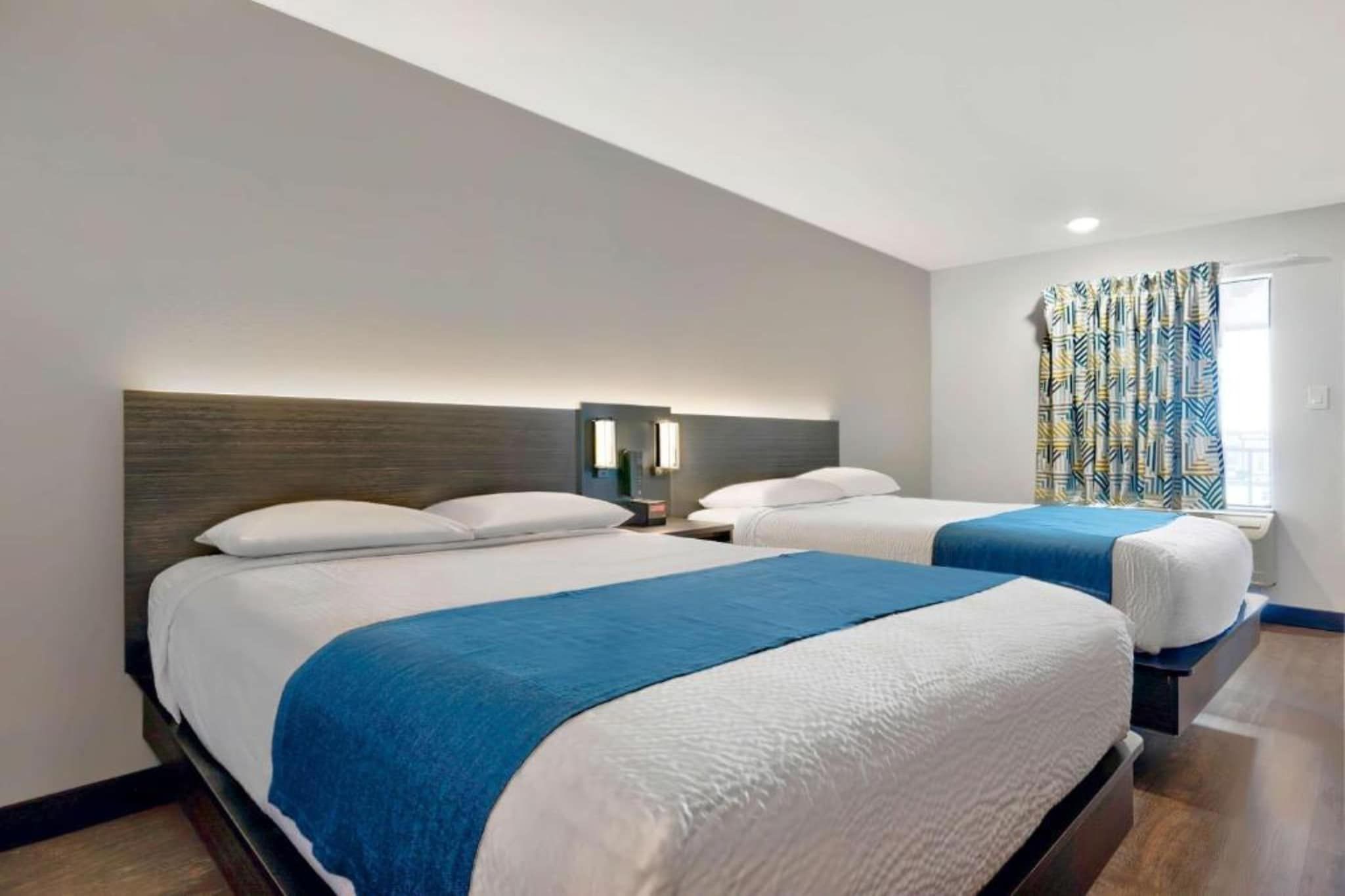 A hotel room with two beds, a nightstand with a lamp, and a window with curtains.