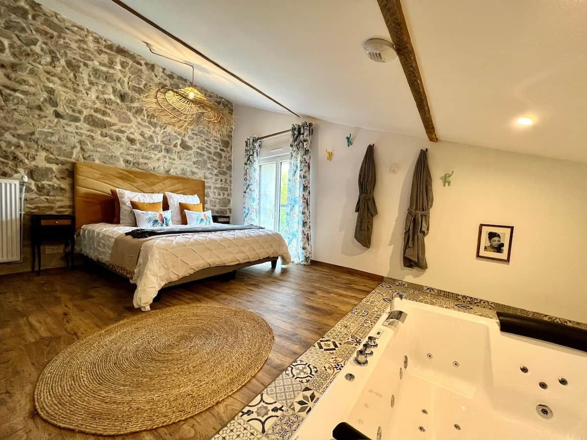 Een slaapkamer met een stenen muur, een bed met houten hoofdeinde, een groot raam met gordijnen, een badkuip en een geweven vloerkleed.