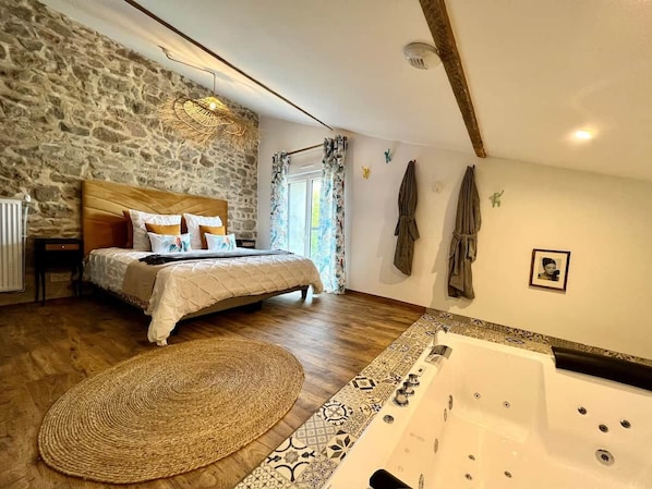 A bedroom with a stone wall, a bed with a wooden headboard, a large window with curtains, a bathtub, and a woven rug.