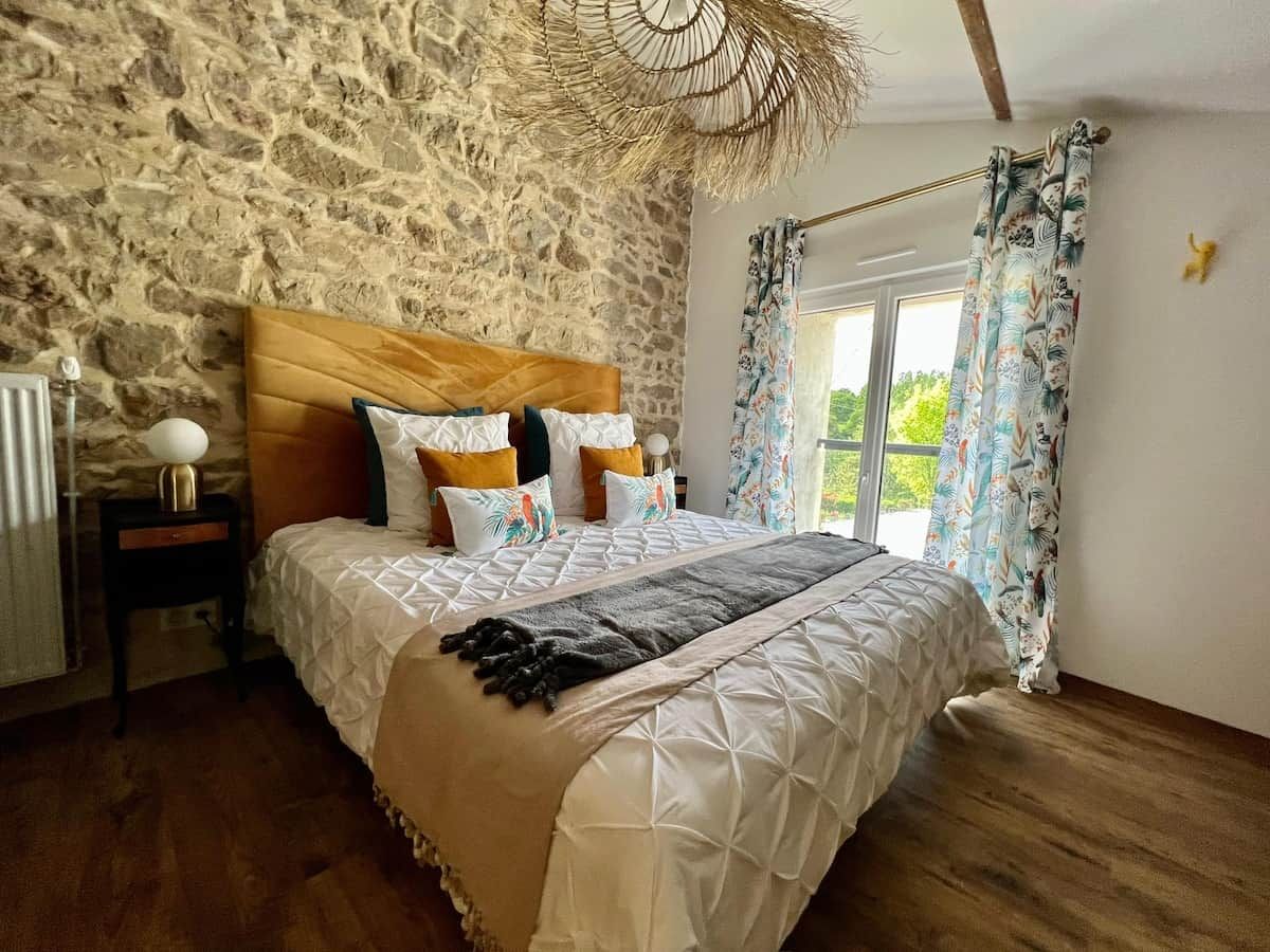 Un dormitorio con pared de piedra, cabecera de madera, cama con ropa blanca, mesita de noche con lámpara y ventana con cortinas.