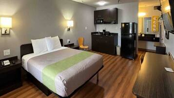 A hotel room with a bed, a desk, a chair, a kitchenette, and a refrigerator.