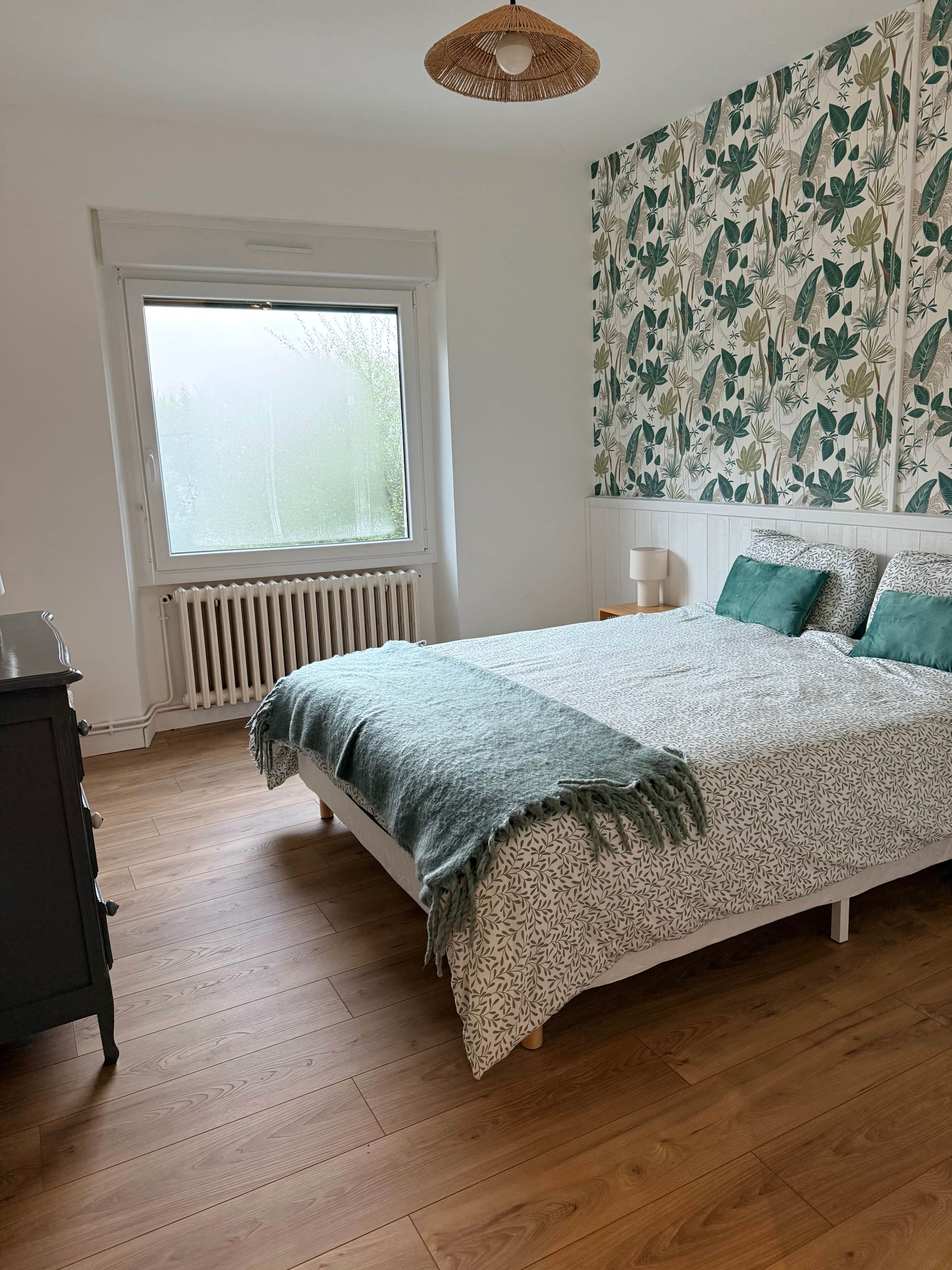 Een slaapkamer met een groot raam, een bed met beddengoed met een patroon, een radiator en een muur met een bladmotief.
