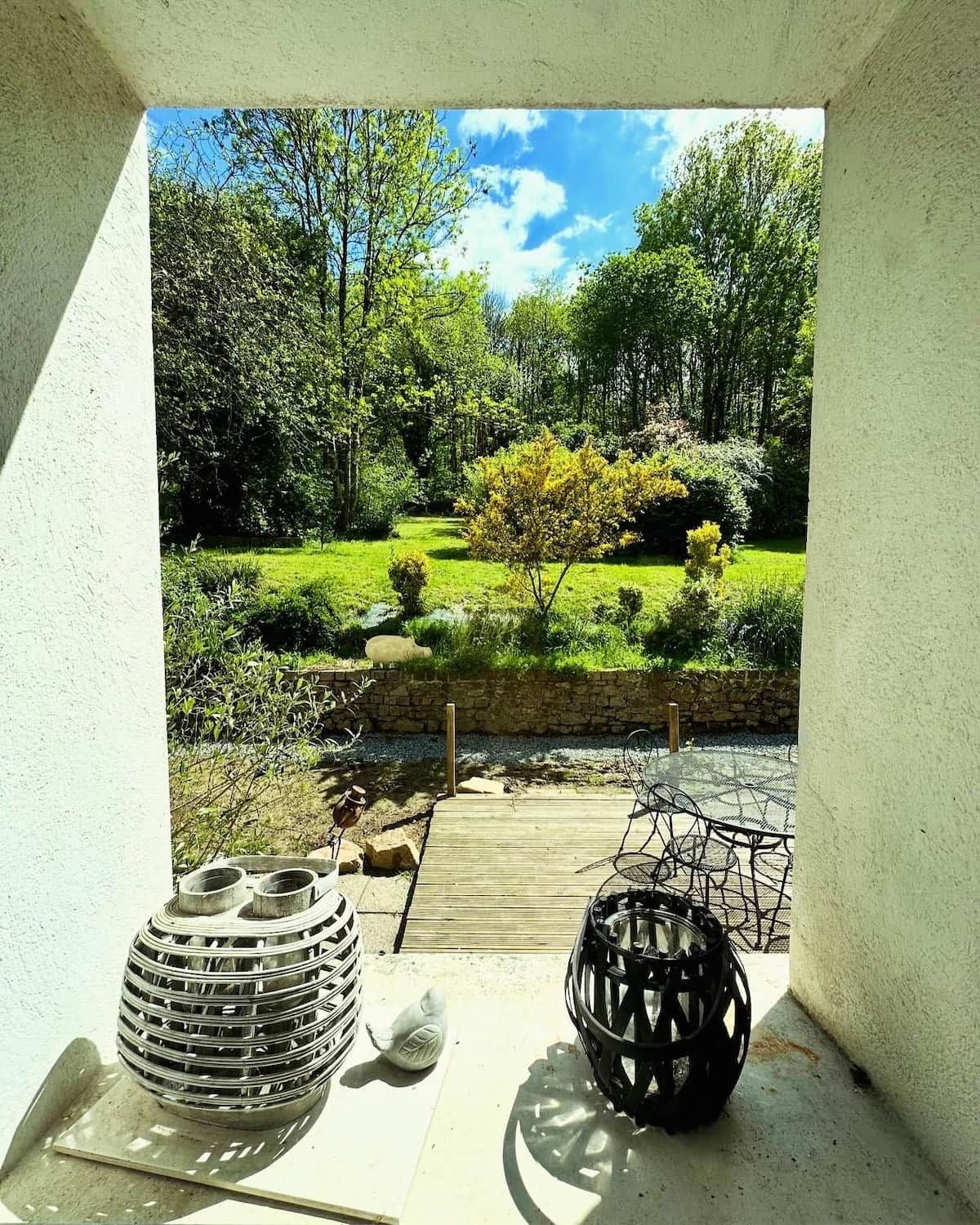 Een terras met tuinmeubilair, een kleine tuin met een vijver en uitzicht op een bebost gebied.