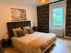 Quarto com cama, mesa de cabeceira, radiador, janela e parede com padrão de folhagem.