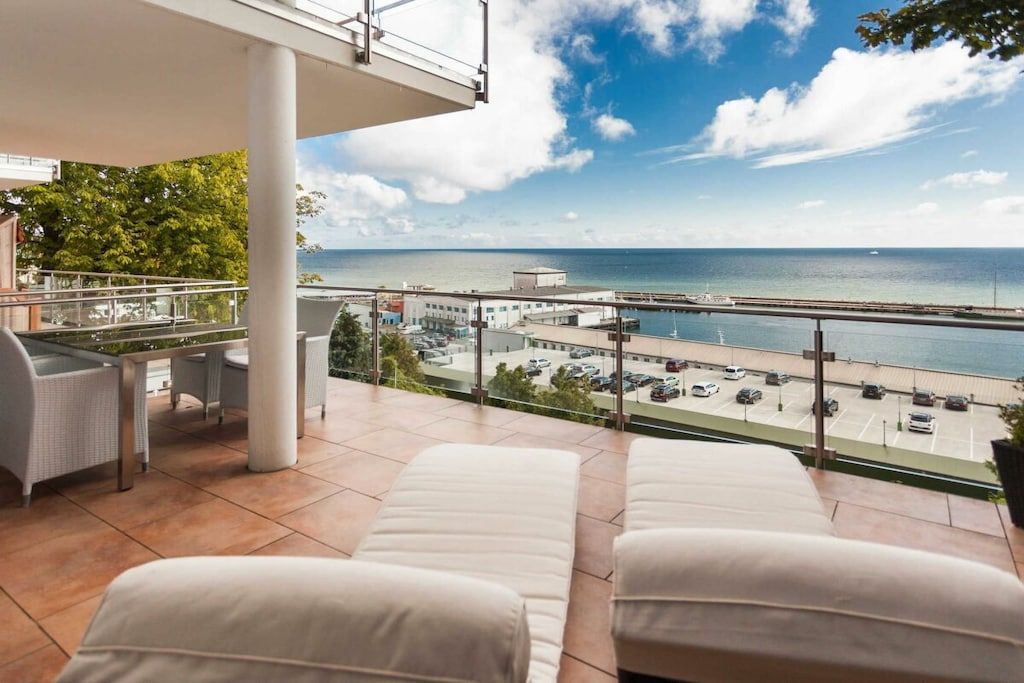 A balcony with two reclining chairs, a table, and a view of the sea and a bridge.