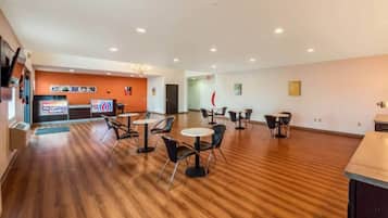 A spacious indoor area with wooden flooring, multiple tables and chairs, a reception desk, and a television screen.
