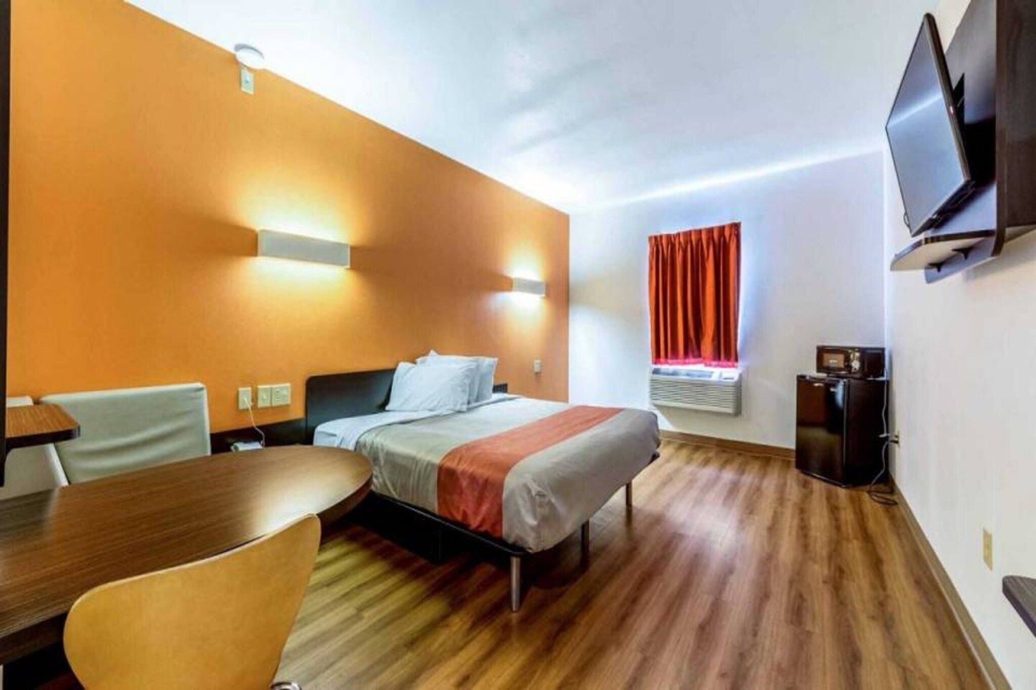 A hotel room with a bed, a desk, a chair, a TV, and an air conditioning unit.