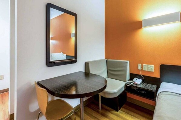 A hotel room with a bed, a bedside table, a chair, a small table, a mirror, and a wall-mounted phone.