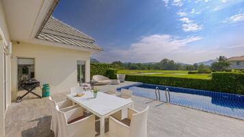 An outdoor patio with a pool, a table, and chairs, overlooking a green landscape.