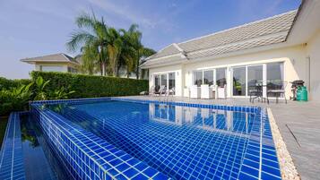 A swimming pool with a tiled edge, a patio area with outdoor furniture, and a building with large windows and a tiled roof.