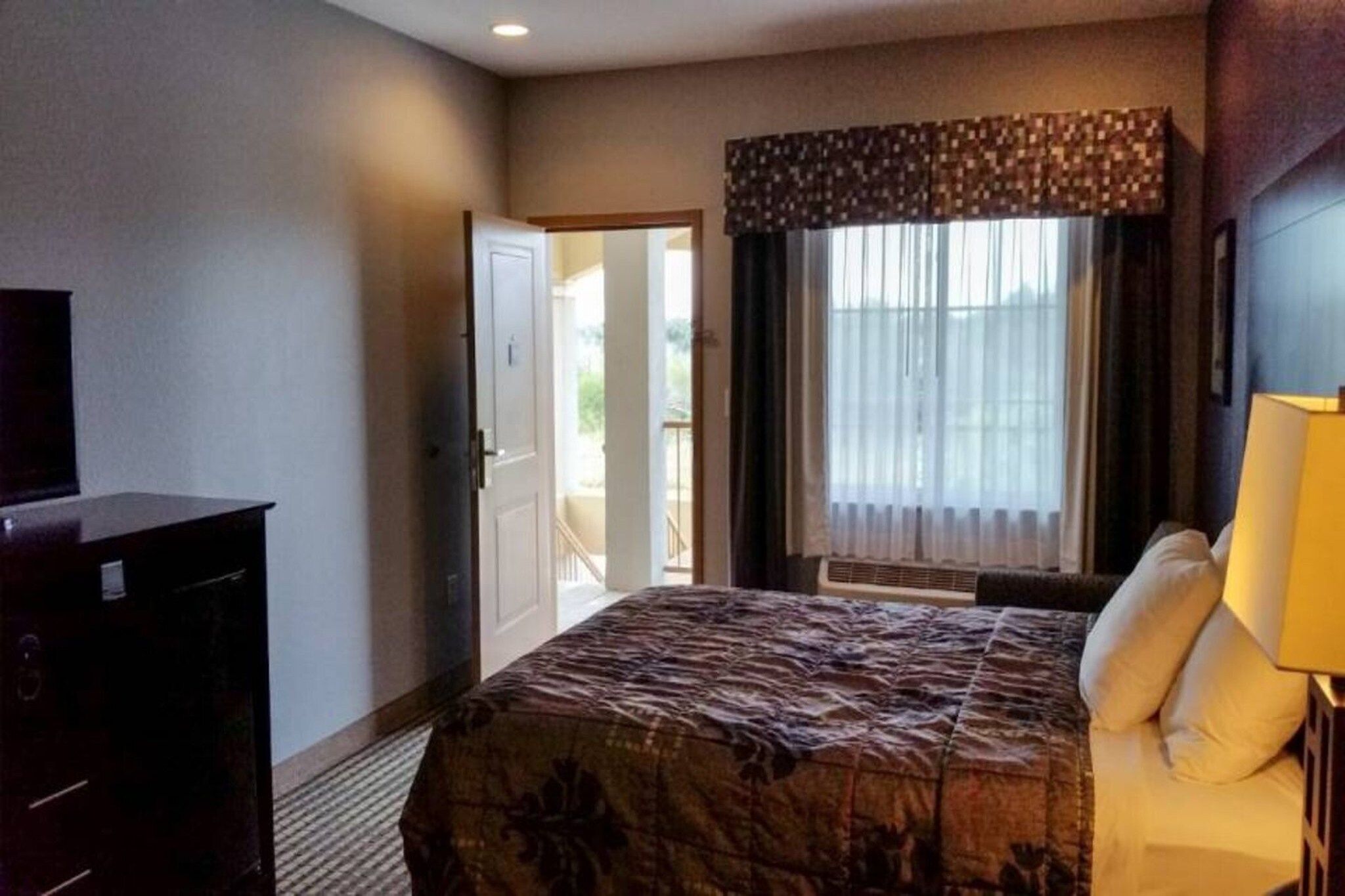 Quarto de hotel com cama, escrivaninha, televisão, janela com cortinas e porta para uma varanda.