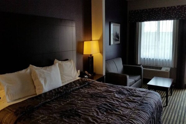 A hotel room with a large bed, a bedside table with a lamp, a small table, a chair, and a window with curtains.