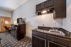 A compact hotel room with a kitchenette, a single bed, a desk with a chair, and a large window.