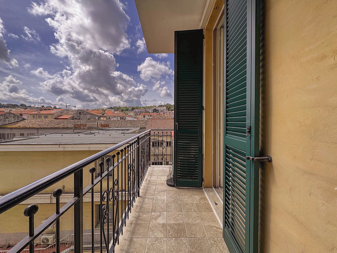 Un balcon avec une balustrade en métal, une porte verte et une vue sur une étendue d’eau et des bâtiments au loin.
