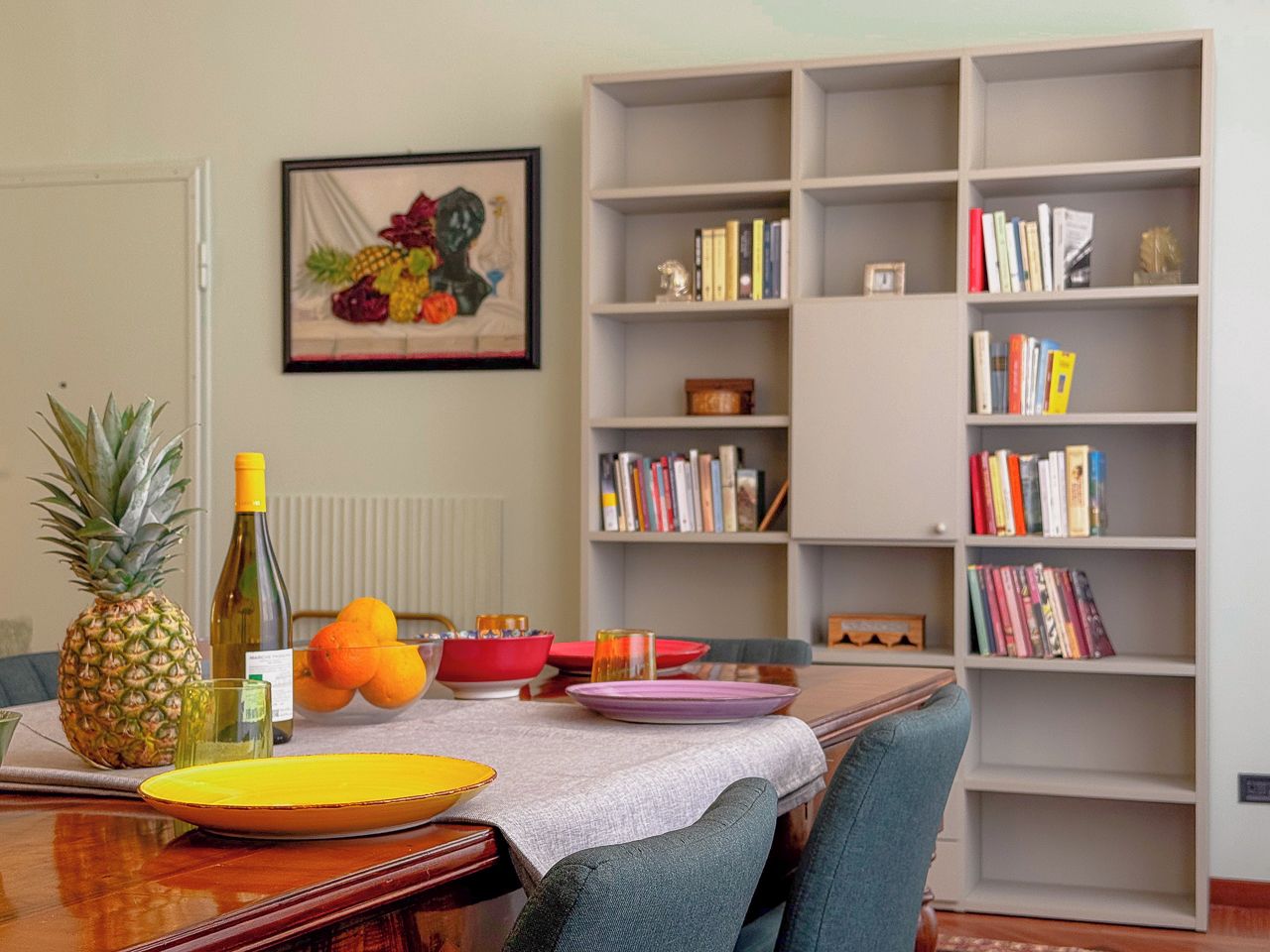 Un comedor con mesa de madera, sillas, una estantería y una obra de arte enmarcada en la pared.