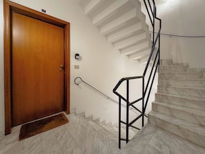 Eine Holztür mit Fußmatte, eine Marmortreppe mit schwarzen Metallgeländern und eine weiße Wand mit Lichtschalter.