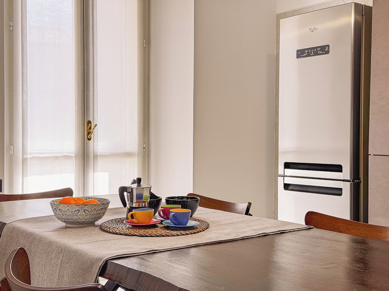 Un comedor con una mesa preparada para dos personas, un cuenco de fruta y una cafetera.