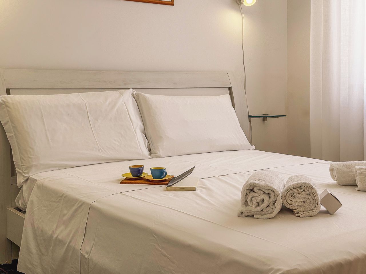 Un lit bien fait, avec du linge de lit blanc, une pile de serviettes enroulées, un livre et deux tasses sur un plateau.