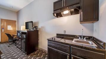 A compact hotel room with a kitchenette, a single bed, a desk with a chair, and a large window.