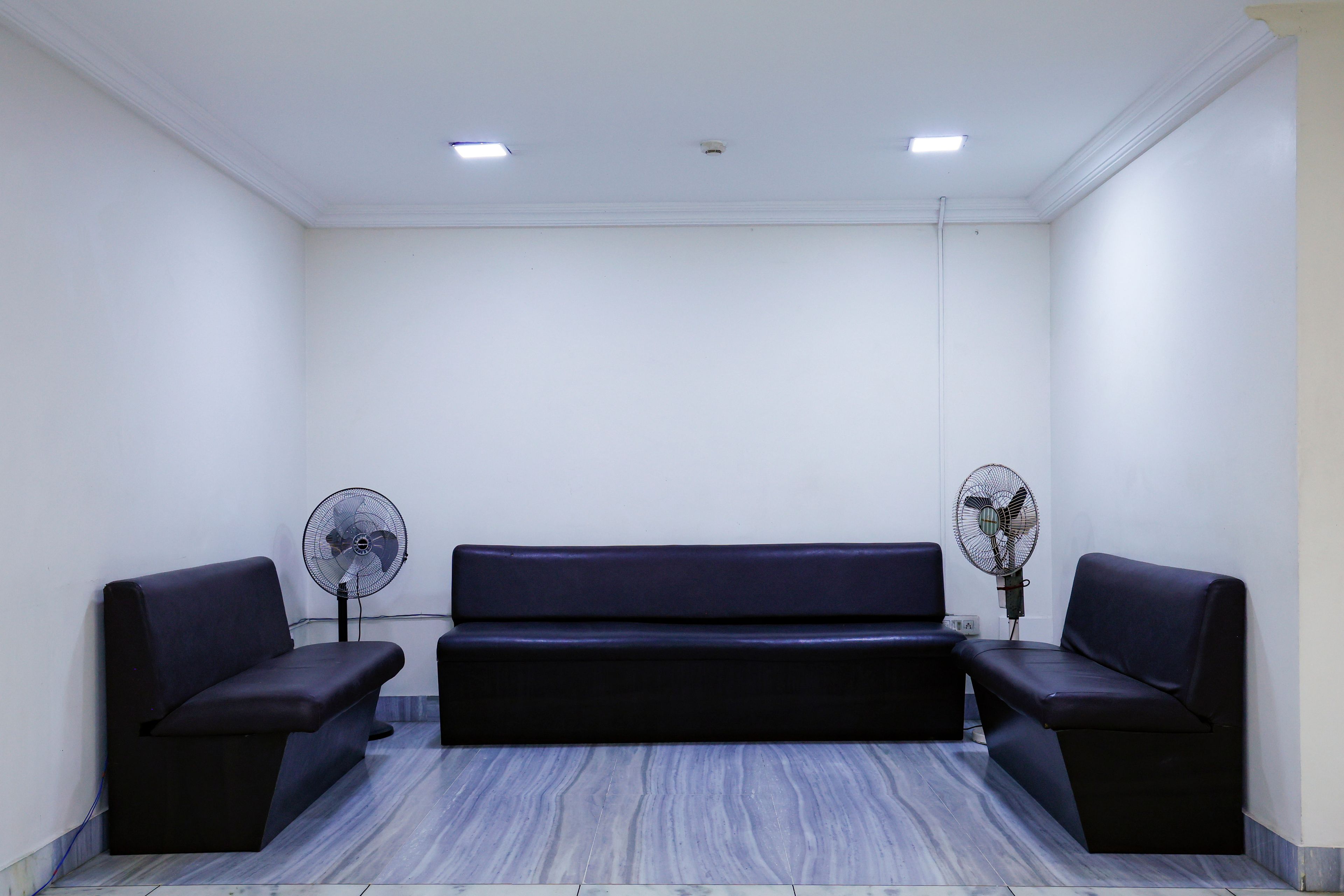 A room with a dark-colored sofa, two standing fans, and a marble-patterned floor.