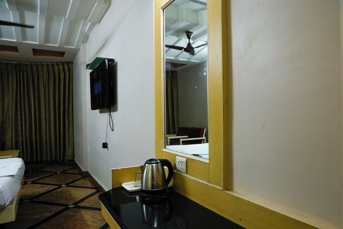 A hotel room with a bed, a desk with a kettle, a television, and a window with a ceiling fan.