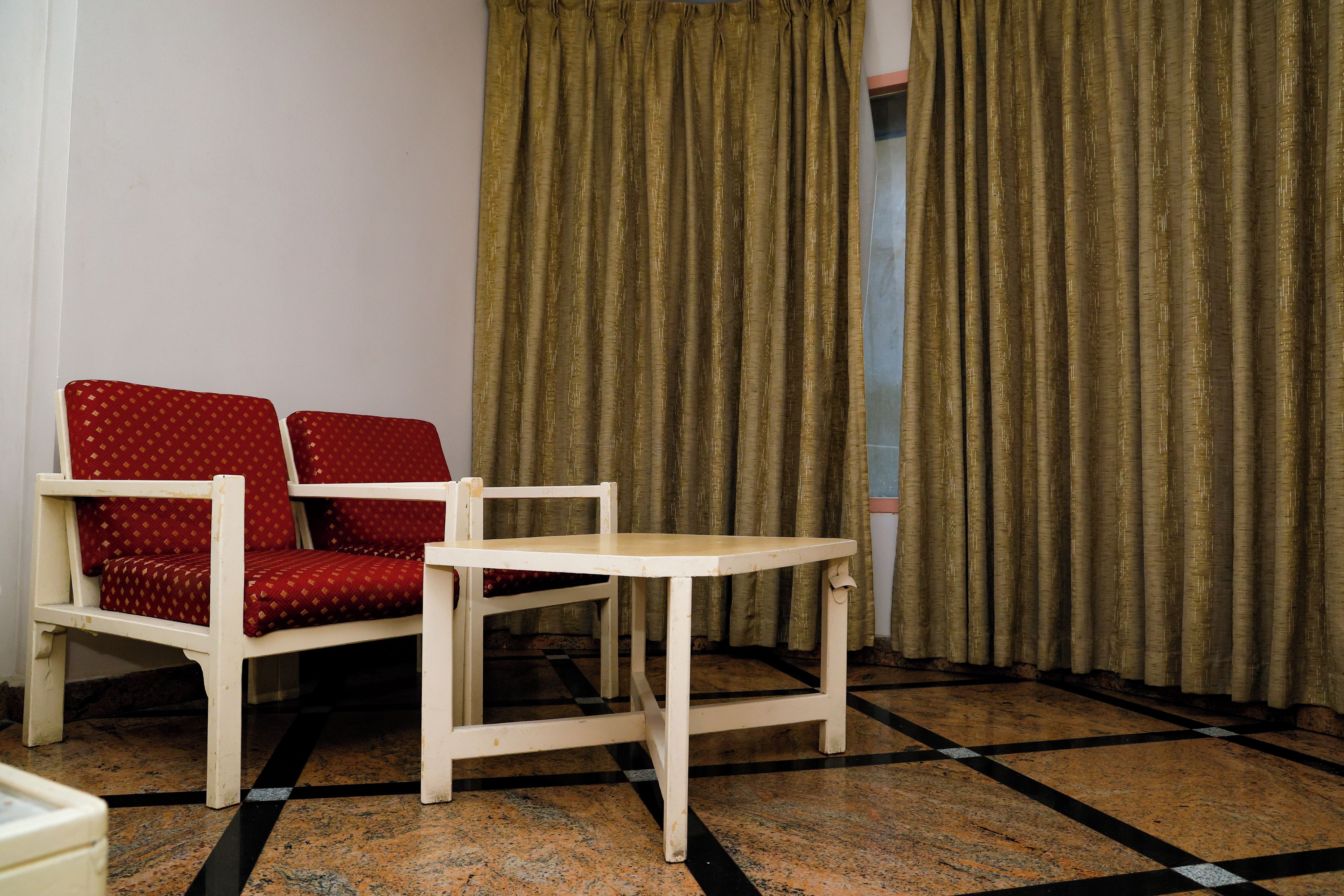 Une pièce avec une chaise blanche et une petite table, un canapé rouge à motifs et une paire de rideaux.