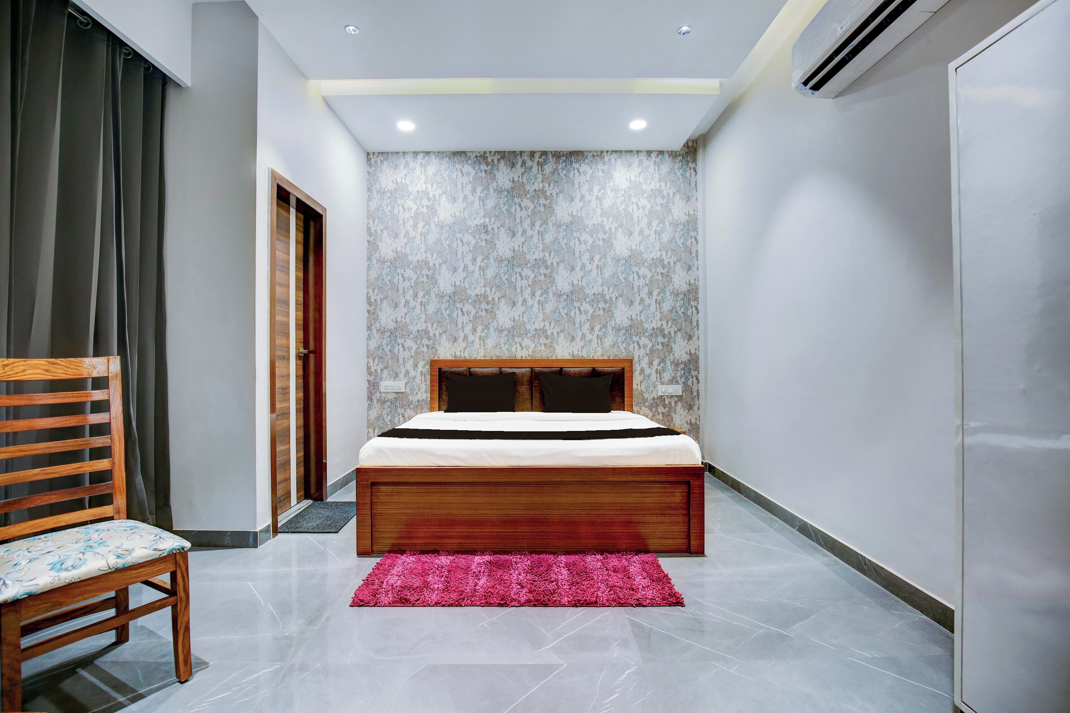 Un dormitorio con cama, una silla, alfombra rosa y pared texturizada.