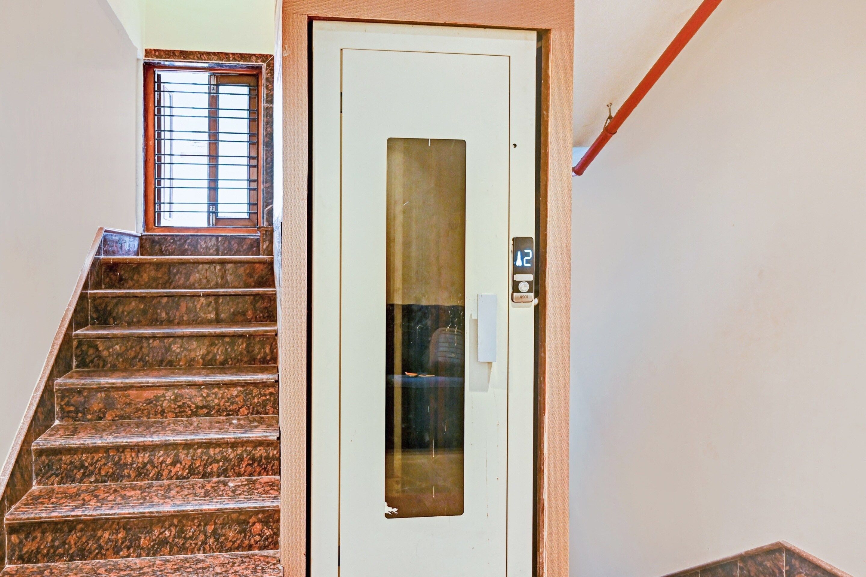 Una escalera lleva a una puerta con una placa de número, una ventana con barrotes y una tubería roja en la pared.