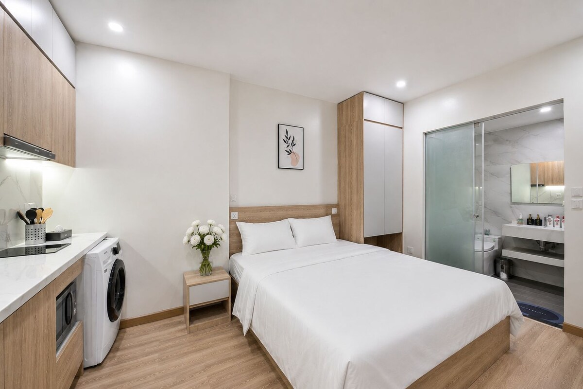 A modern hotel room with a bed, a washing machine, a small table with a vase of flowers, and a bathroom with a glass shower.