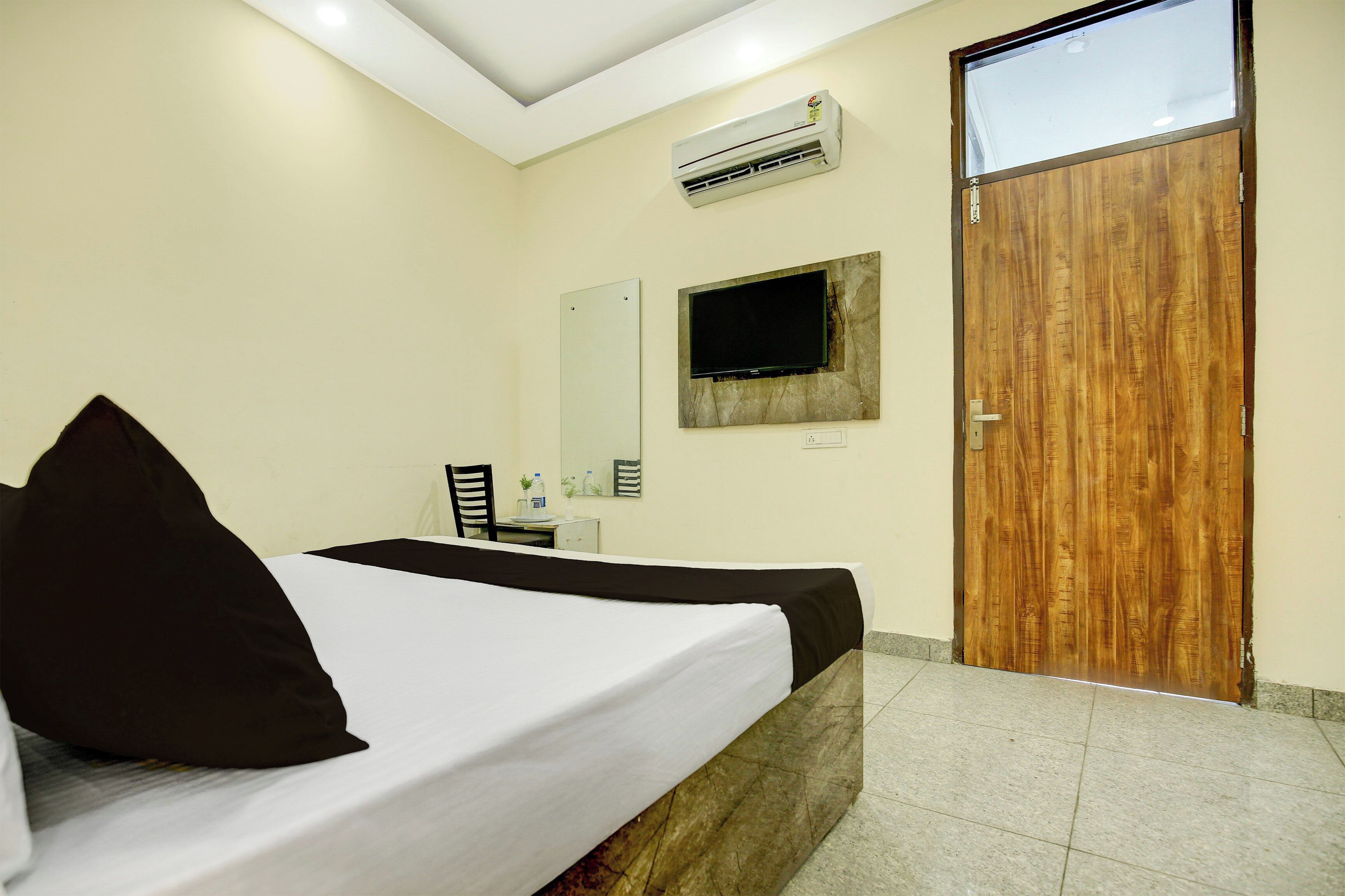 A hotel room with a bed, a flat-screen TV mounted on the wall, an air conditioning unit, and a wooden door.