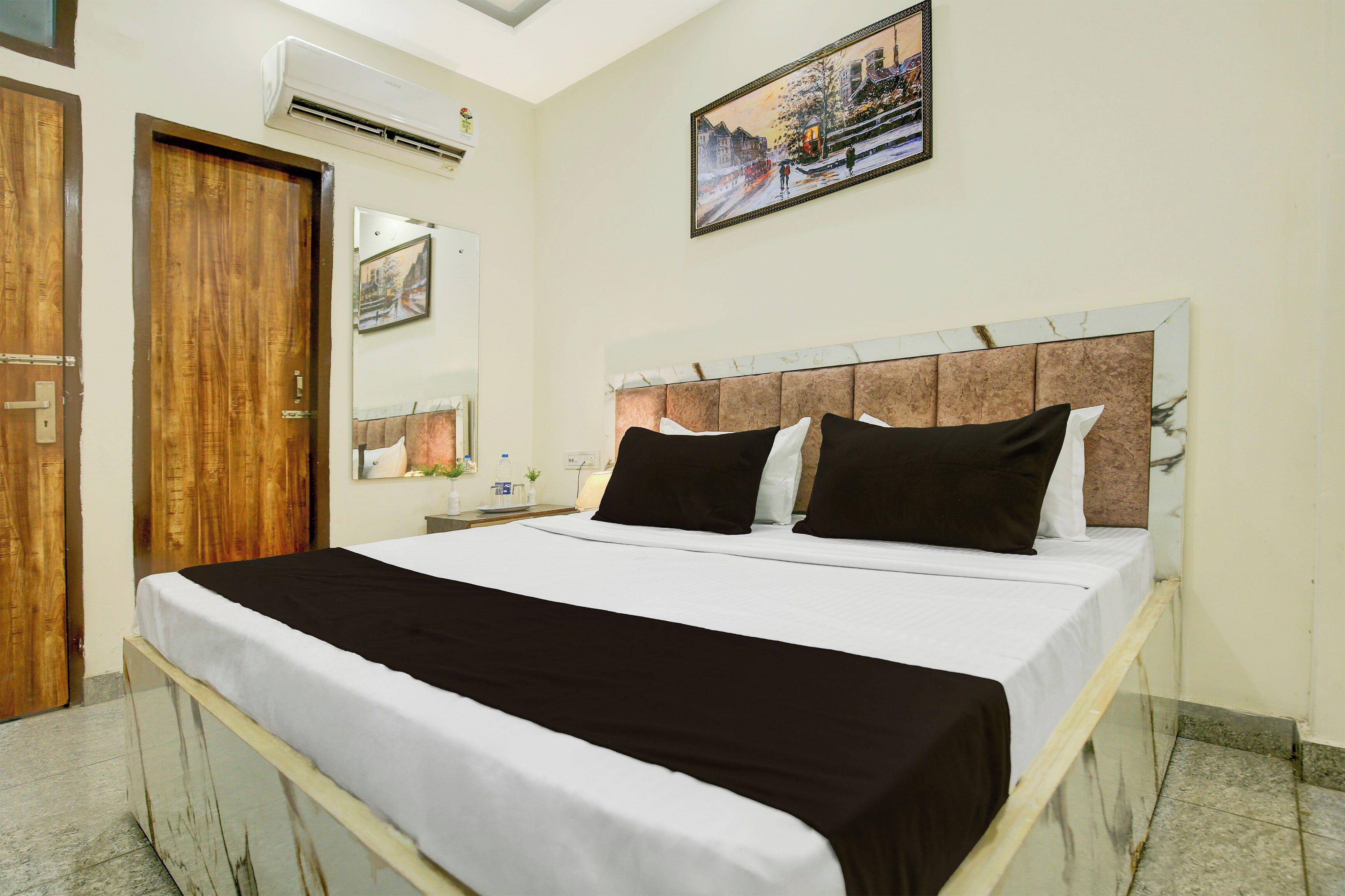 A hotel room with a bed, a wooden door, an air conditioning unit, a framed picture on the wall, and a marble floor.