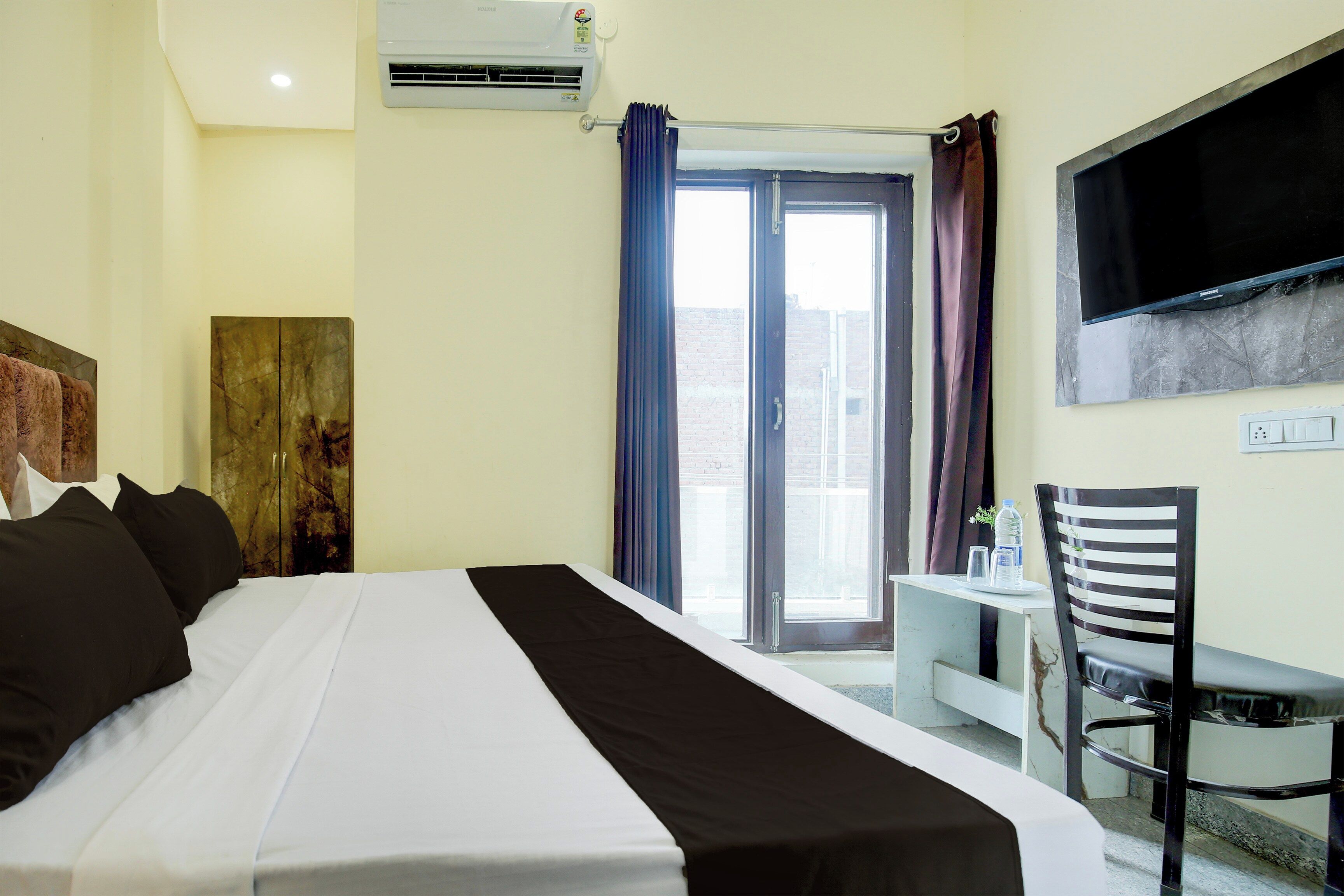 A hotel room with a bed, a TV mounted on the wall, a desk with a chair, and a window with curtains.