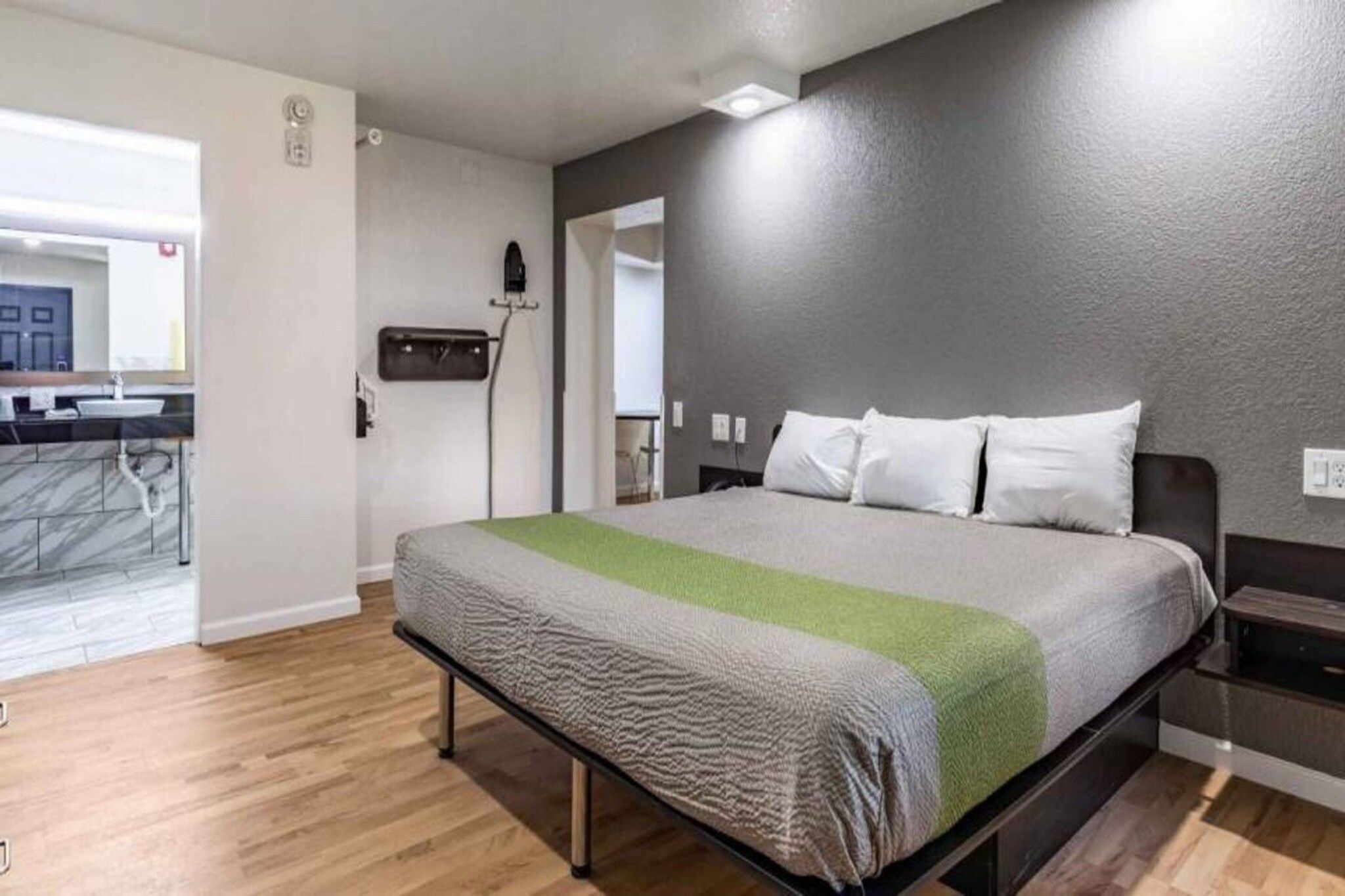 A hotel room with a bed, a nightstand, a wall-mounted heater, and a view of the bathroom.