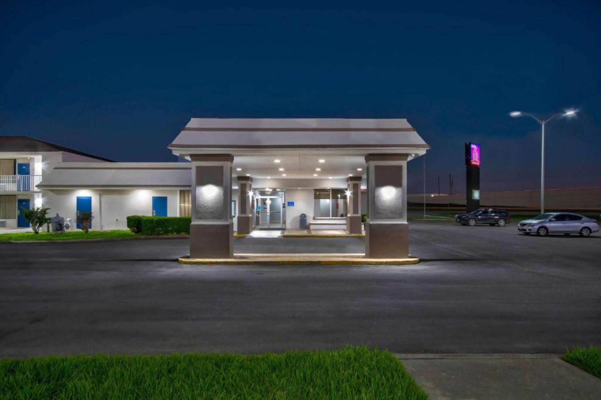 A hotel entrance with a covered waiting area, a parking lot, and a building with a neon sign in the background.