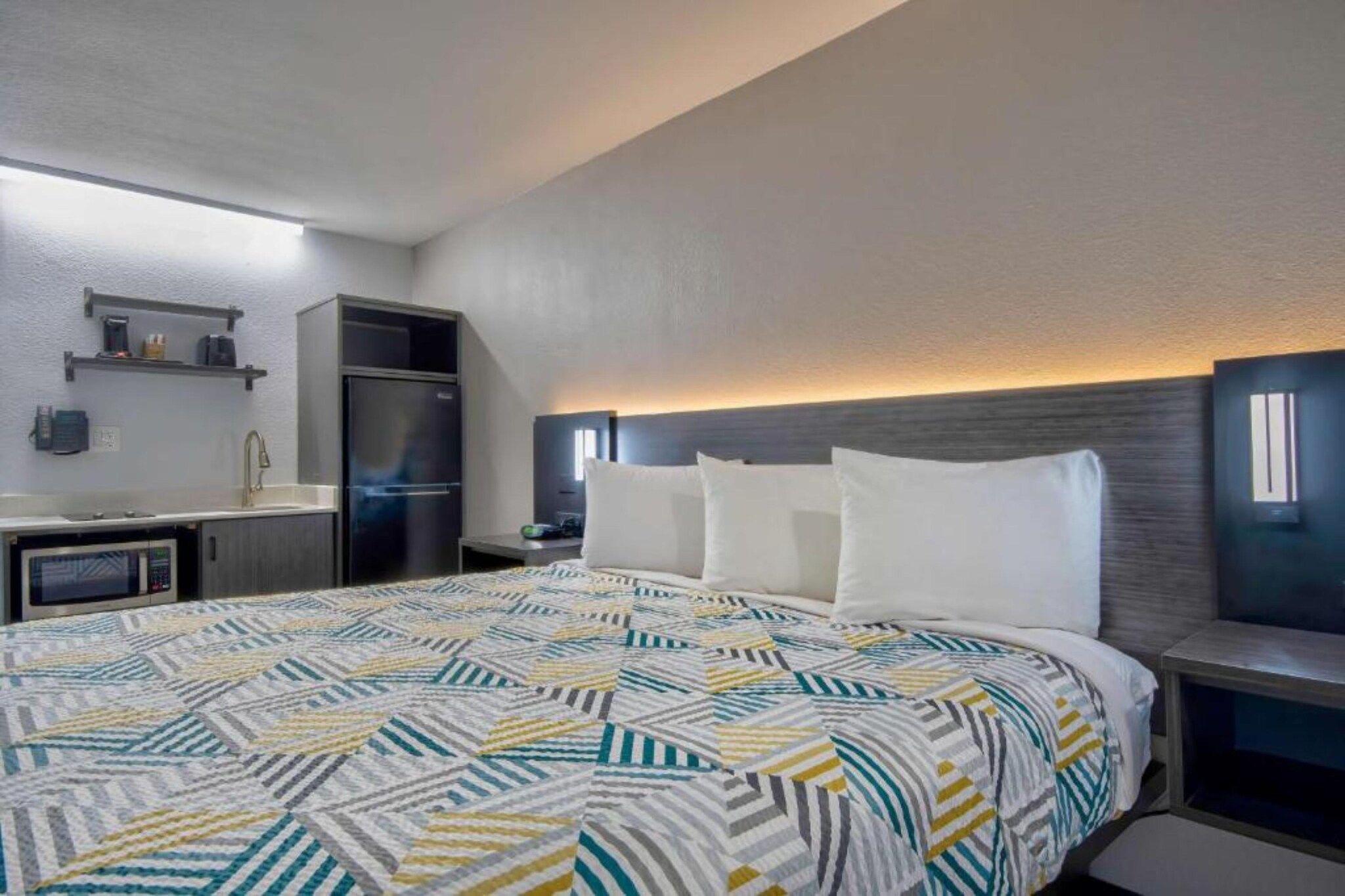 A hotel room with a bed, a kitchenette, a microwave, a refrigerator, and a nightstand with a lamp.
