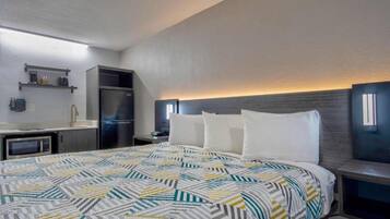 A hotel room with a bed, a kitchenette, a microwave, a refrigerator, and a nightstand with a lamp.