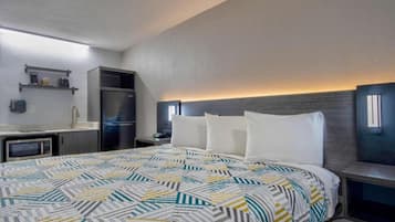A hotel room with a bed, a kitchenette, a microwave, a refrigerator, and a nightstand with a lamp.