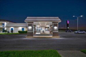 A hotel entrance with a covered waiting area, a parking lot, and a building with a neon sign in the background. - Spacious 1 King Bed | Smk, Kitchen (Rosenberg)