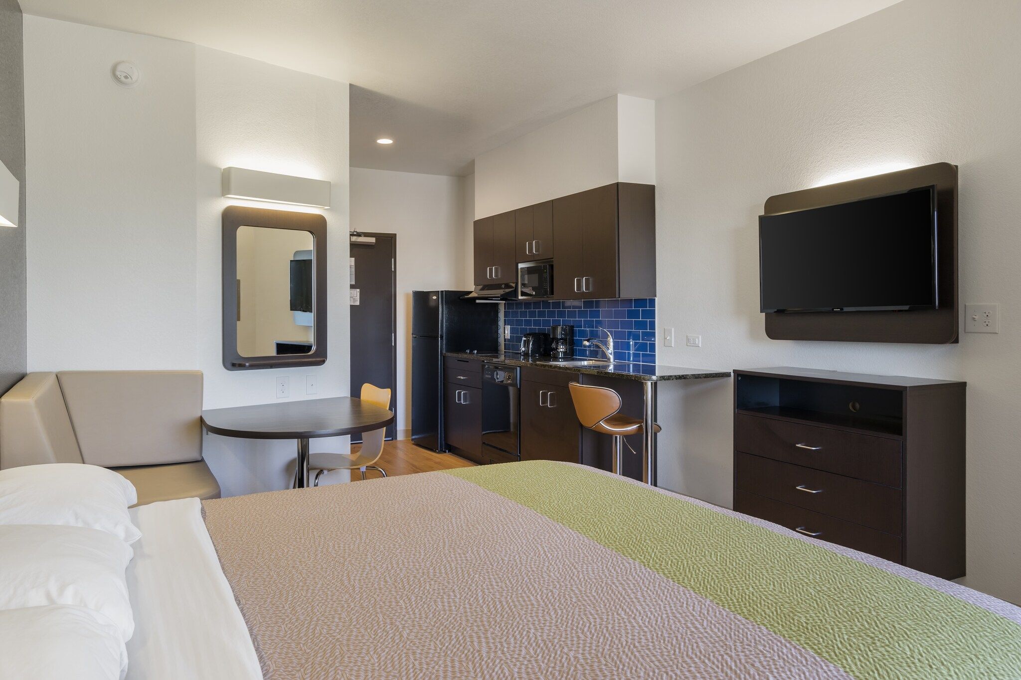 A hotel room with a bed, a desk, a chair, a kitchenette, and a TV.