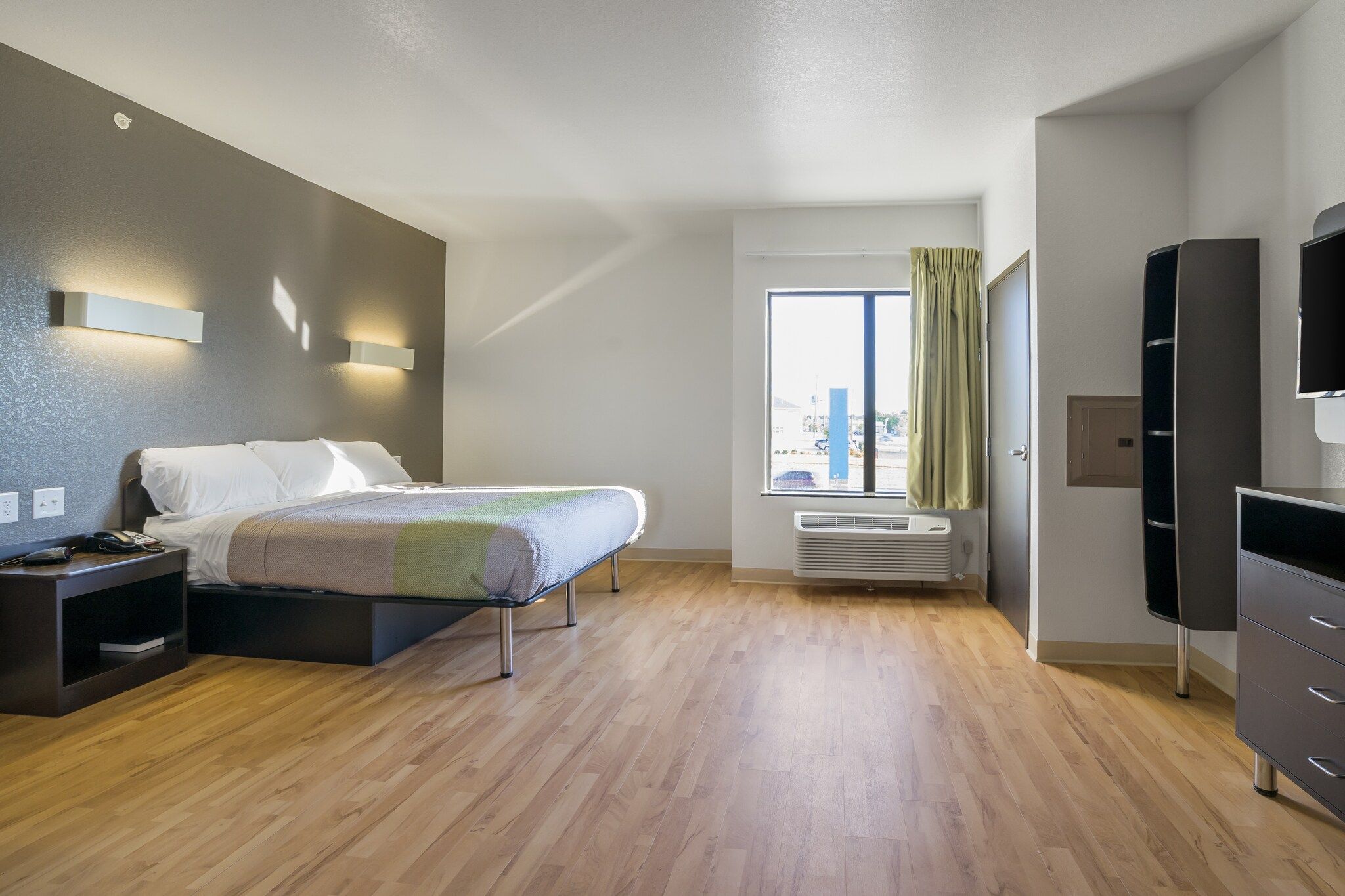 A modern hotel room with a bed, bedside table, TV, wardrobe, and a window with curtains.