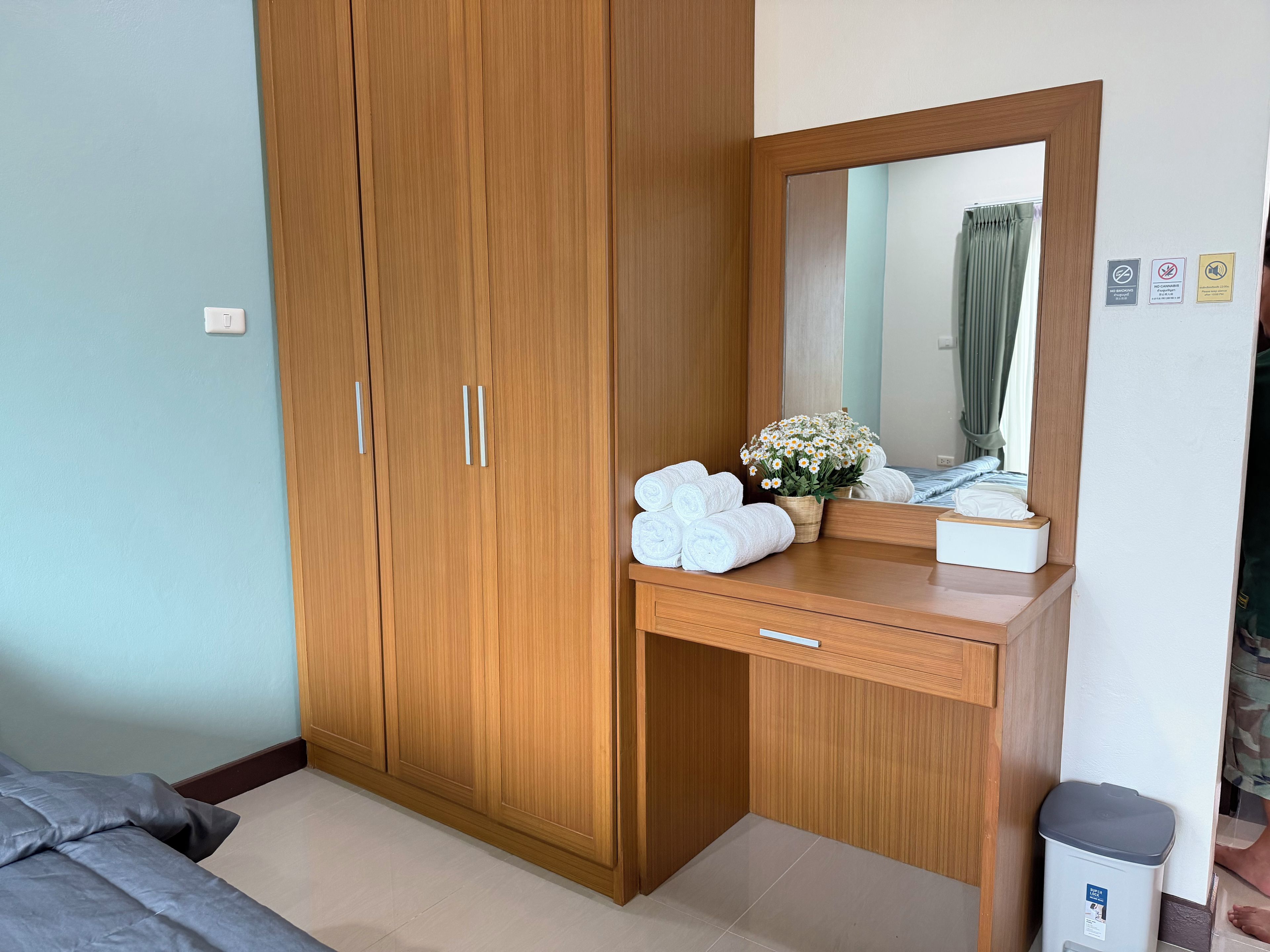 A bedroom with a wooden wardrobe, a bedside table with towels, a mirror, and a trash bin.