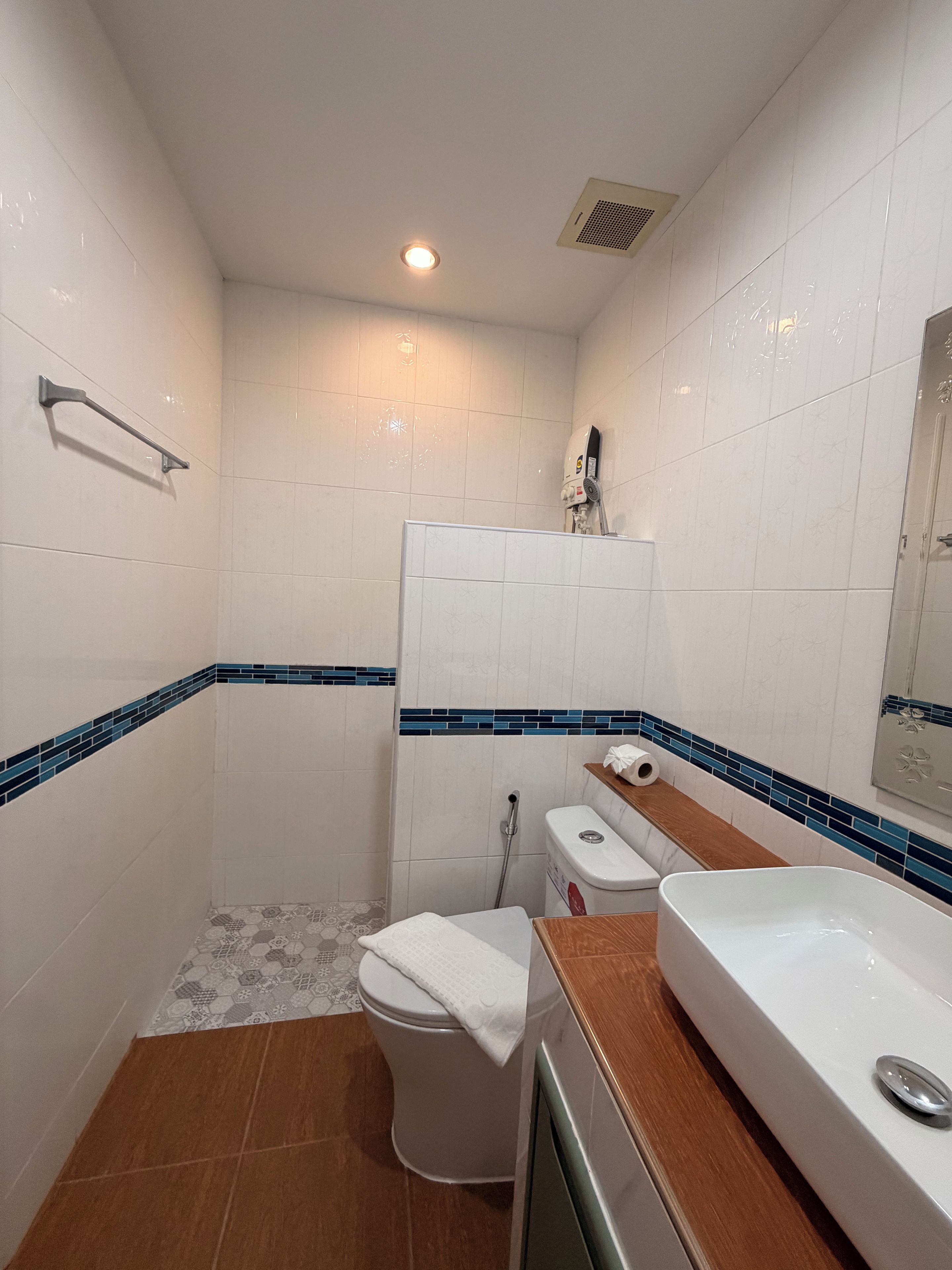 A small bathroom with white tiles, a wooden vanity, a toilet, and a shower area.