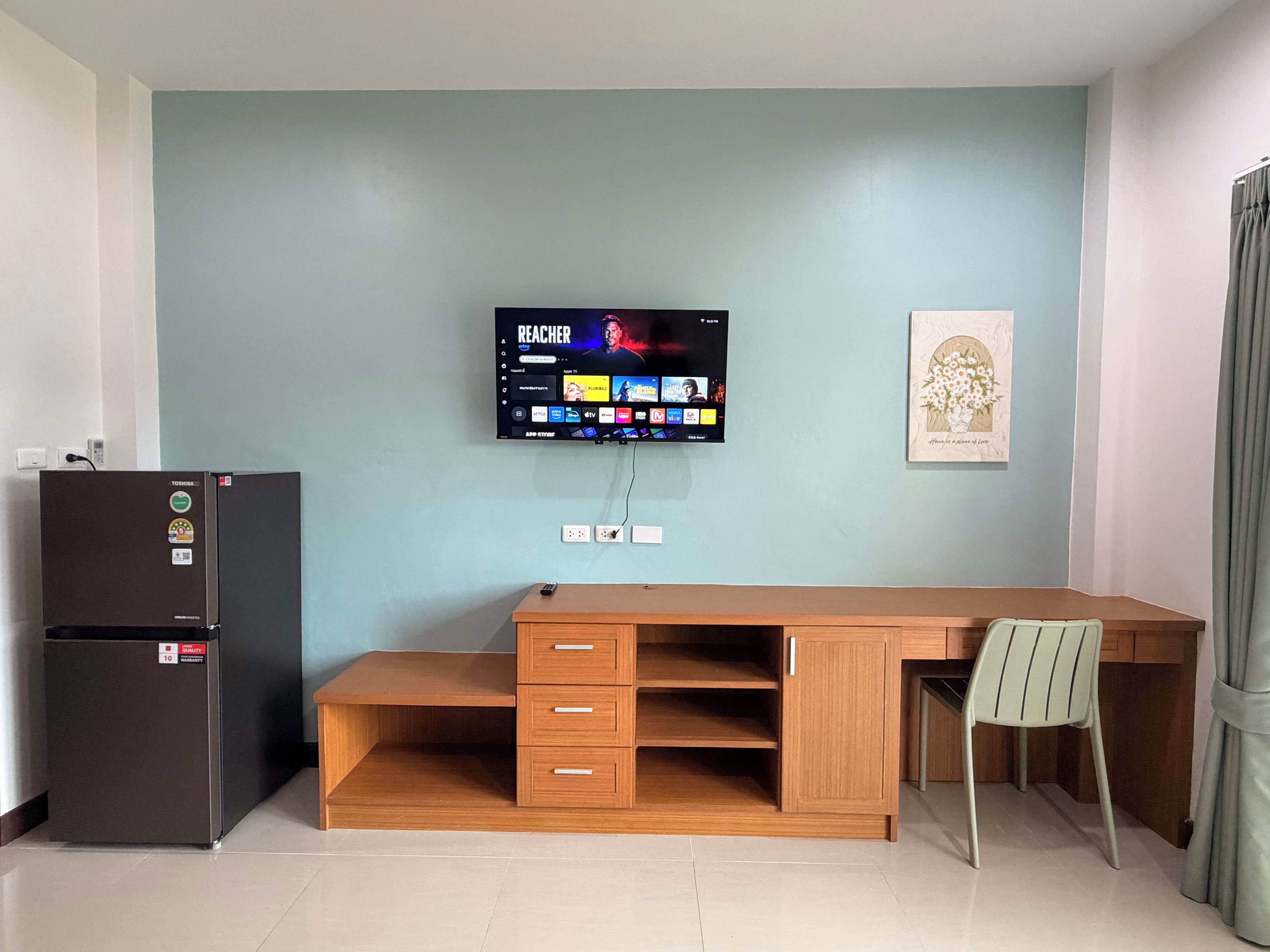 A room with a wooden desk, a television mounted on the wall, a refrigerator, and a chair.