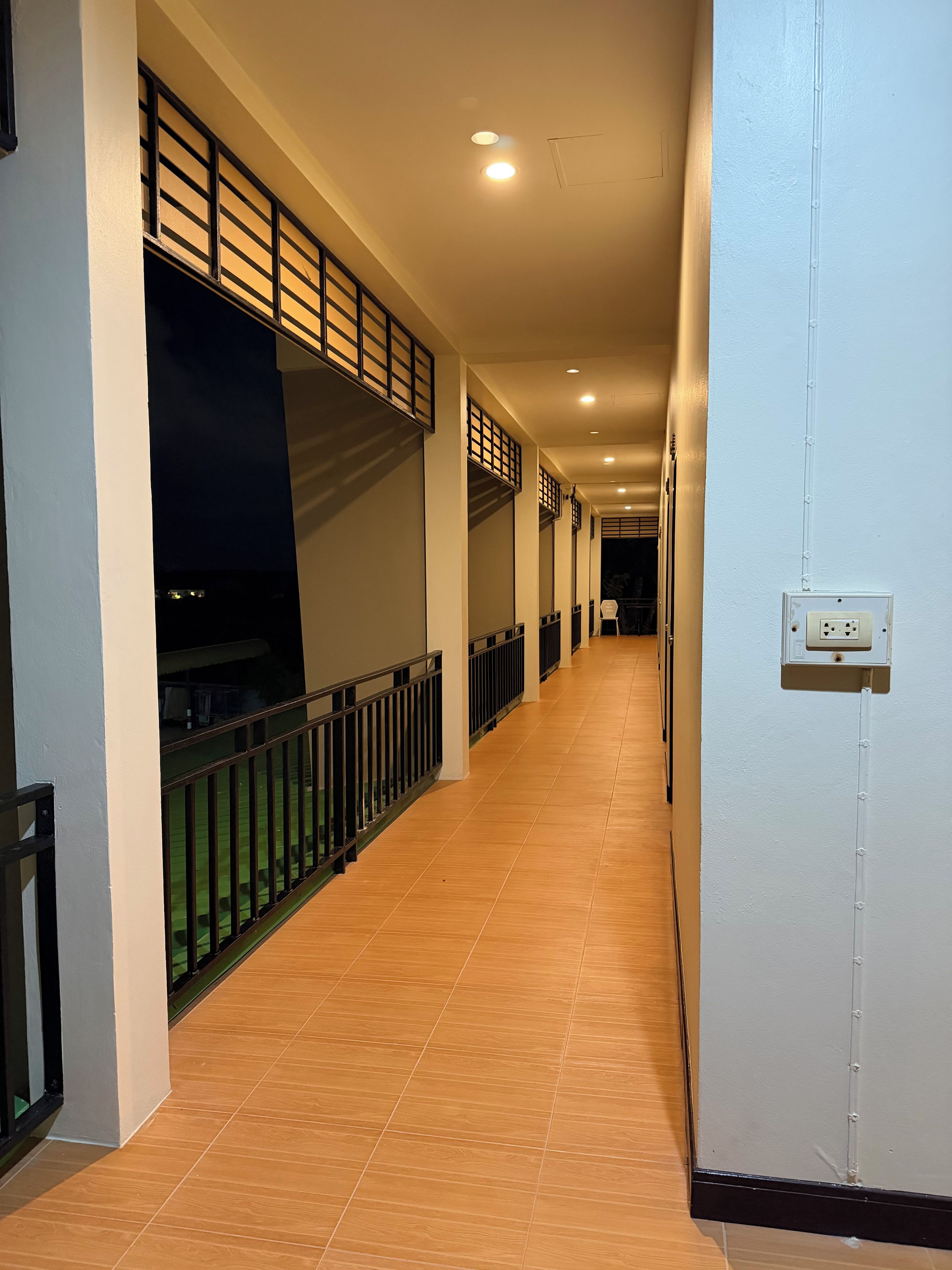A corridor with a tiled floor, a railing on one side, and a wall-mounted electrical outlet on the other.