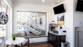 A modern hotel room with a large window, a flat-screen TV, a coffee machine, and a small table with a chair.