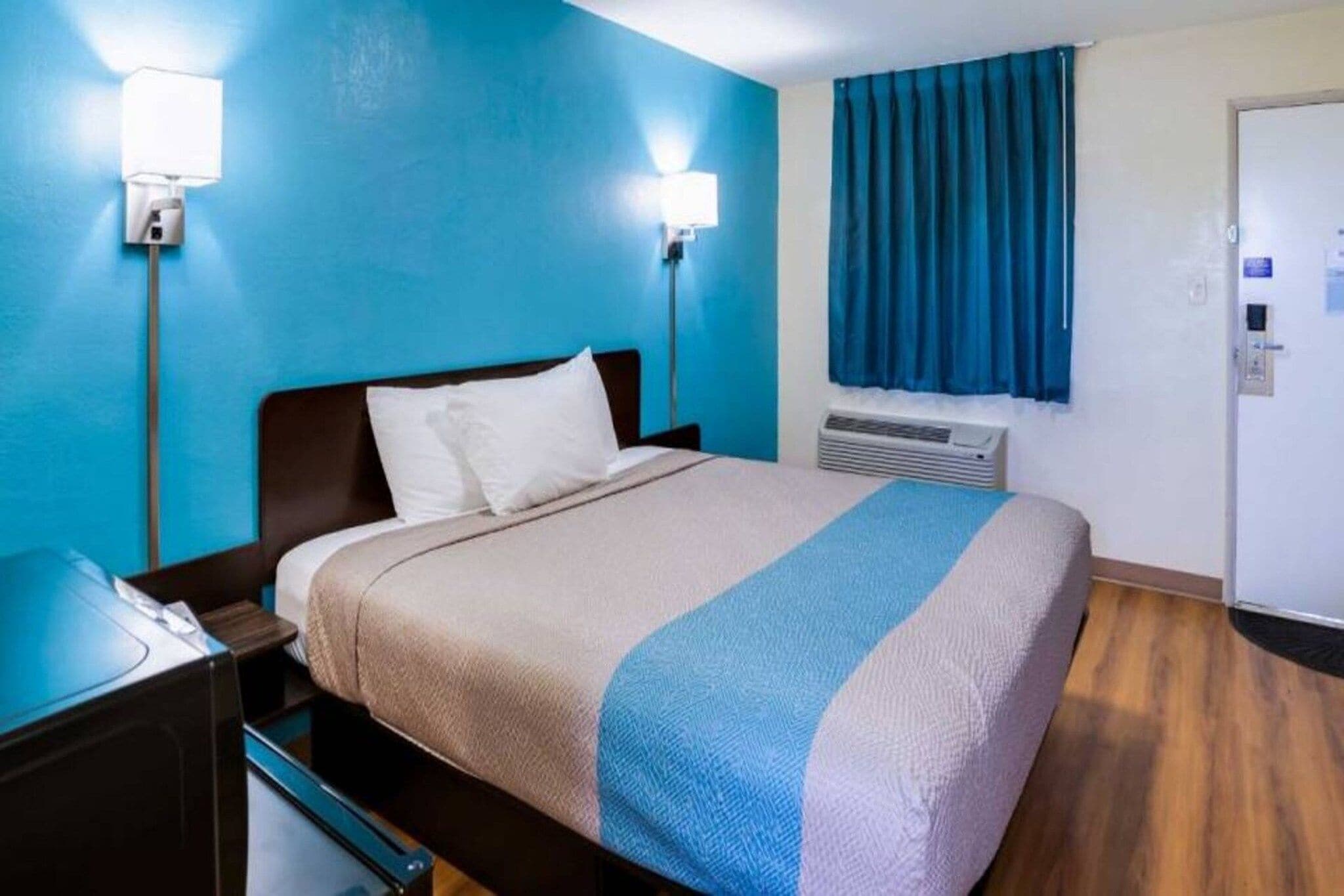 A hotel room with a bed, nightstand, wall-mounted lamp, air conditioning unit, and a window with curtains.